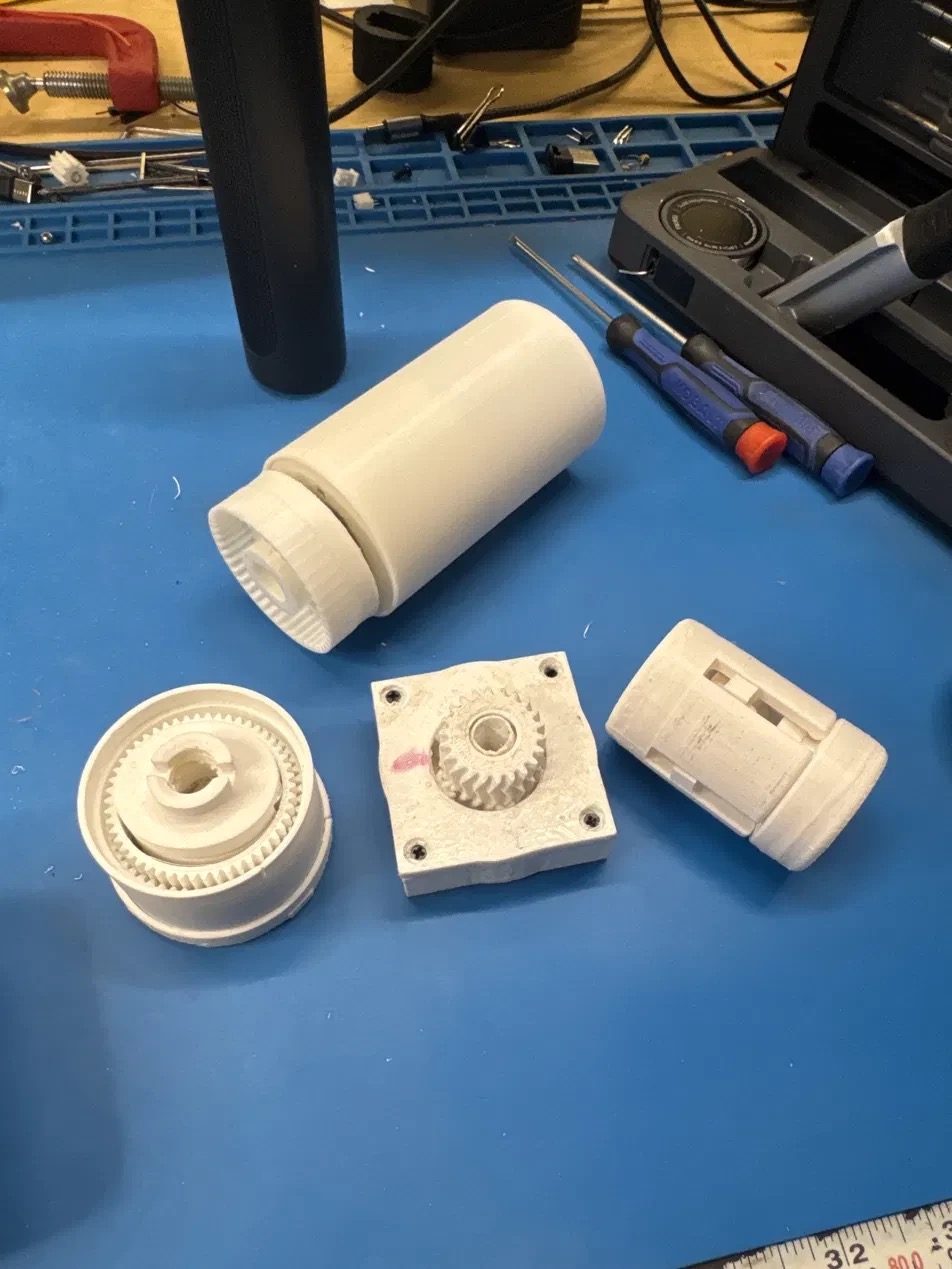 Four printed gearbox prototypes laid out on the blue bench mat — a cylindrical cycloidal housing, a ring-gear stage, a square planetary block, and a split shell